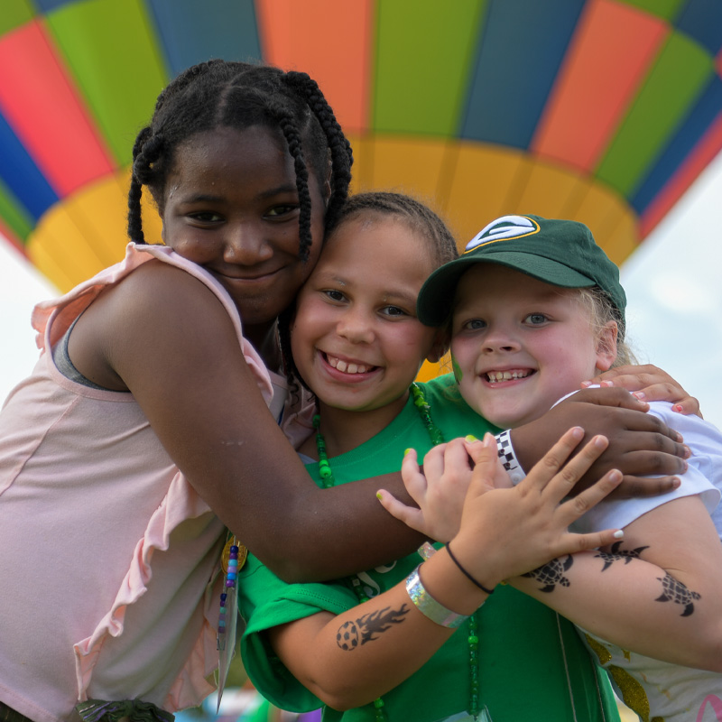 Three girls with skin colors hugging and smiling in front of hot air balloon.