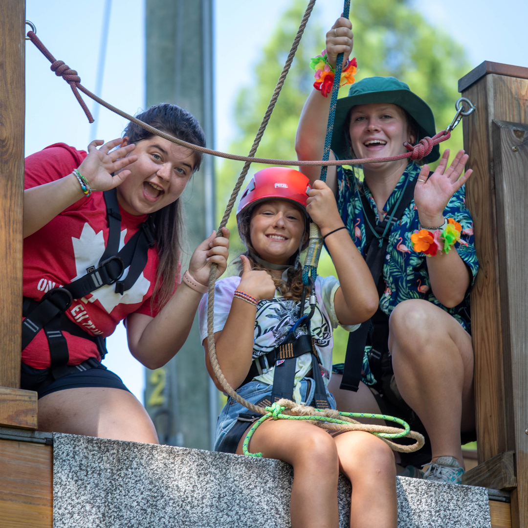 Counselors and camper waving on zipline deck.