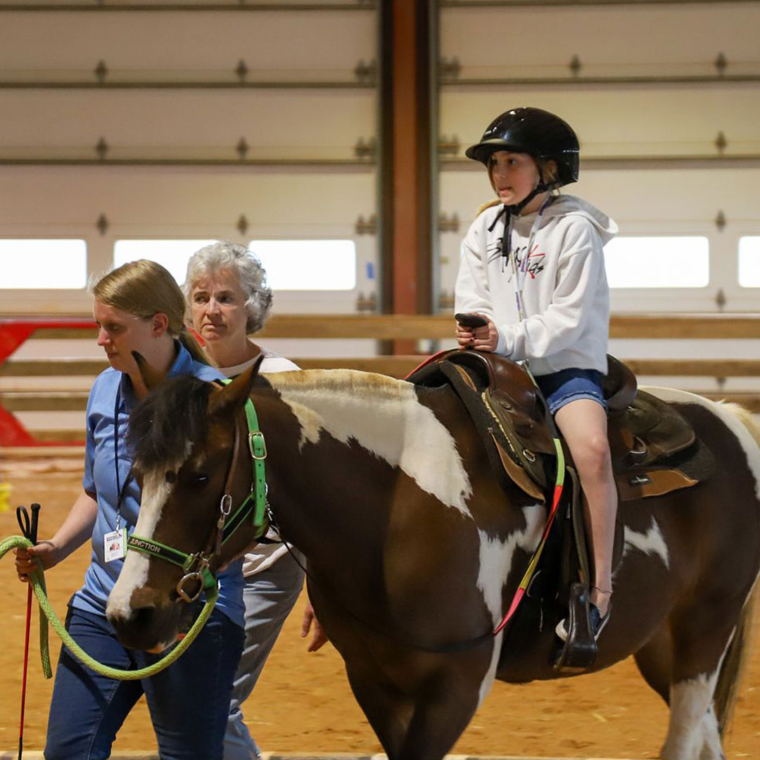 A camper rides a horse with a barn staff member and volunteer leading the horse.