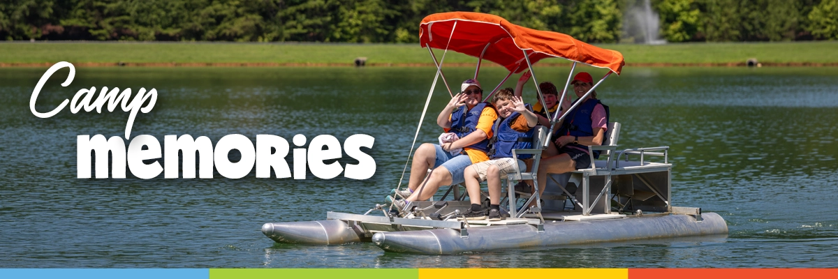Camp Memories Banner 1200x400 Campers and counselors wearing life jackets wave from a small pontoon boat on the camp lake on a sunny day at Victory Junction.