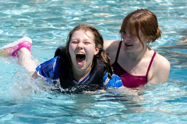 Camp Memories Banner Image 1 600x400 Camper laughing in the pool while a counselor supports them in the water at Victory Junction.