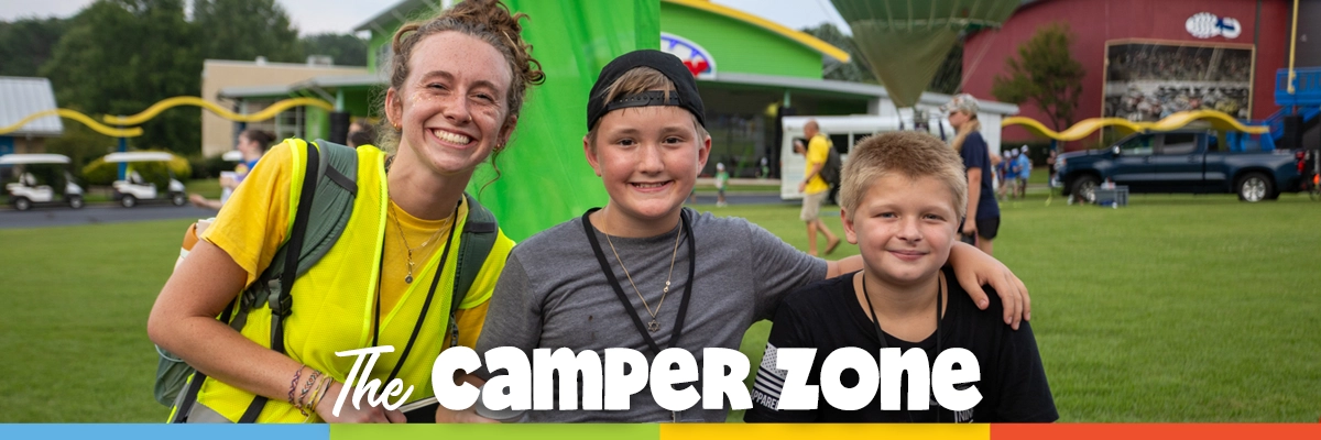 A counselor and two young campers smiling together at Victory Junction, standing on the lawn in front of colorful camp buildings.