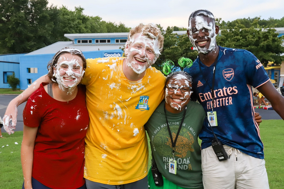 Camp counselors with whipped cream on their faces laugh together outside a building during a messy camp activity.