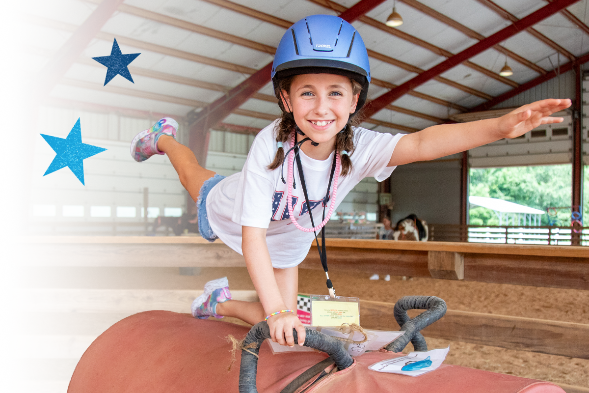 Young camper wearing a helmet balances with arms outstretched on a riding barrel activity in an indoor arena, smiling with confidence.