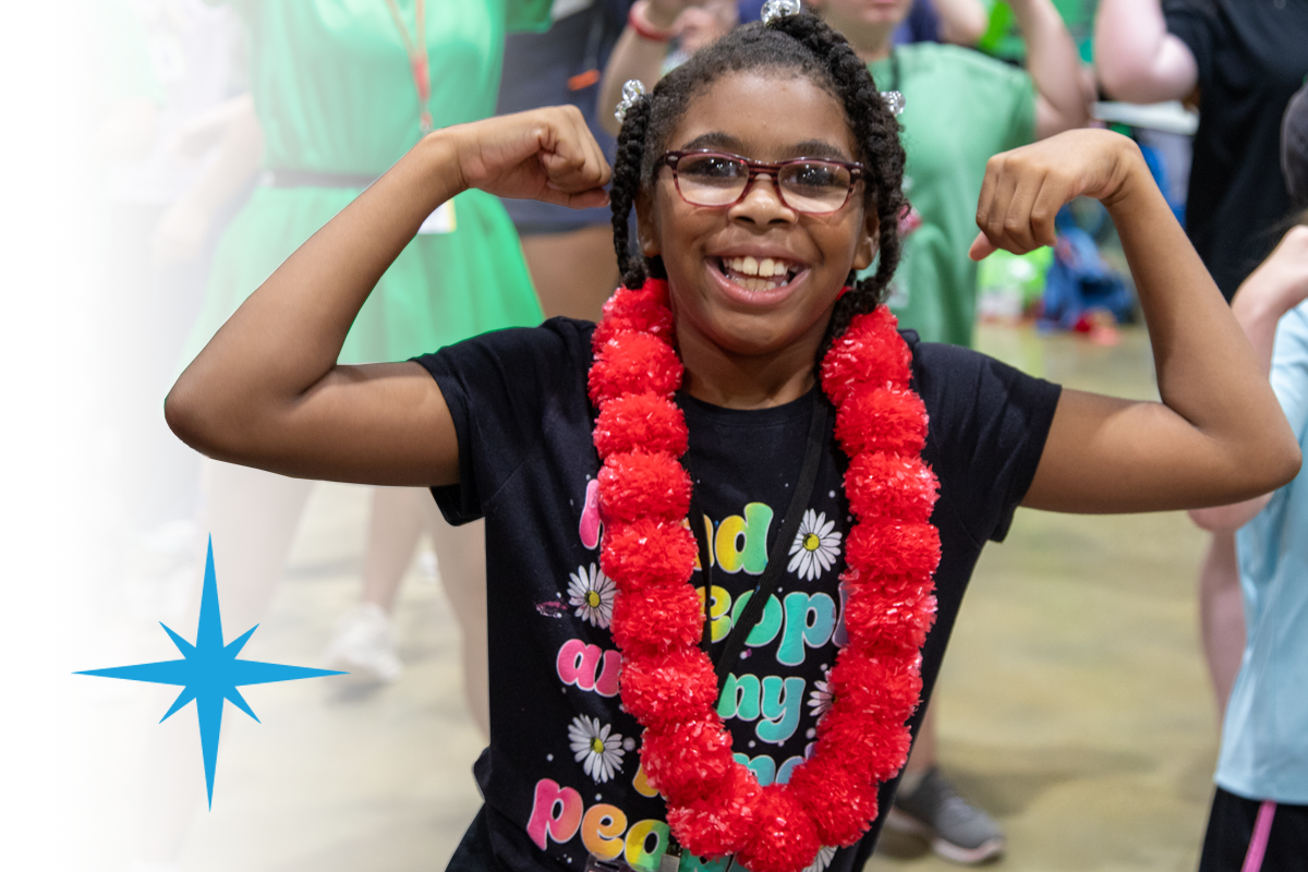 Camper wearing glasses and a red lei smiles and flexes their arms during a dance party, showing joy and confidence.