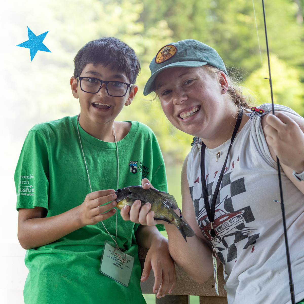 Camper and counselor smile while holding a freshly caught fish by a lakeside, celebrating a shared outdoor experience.
