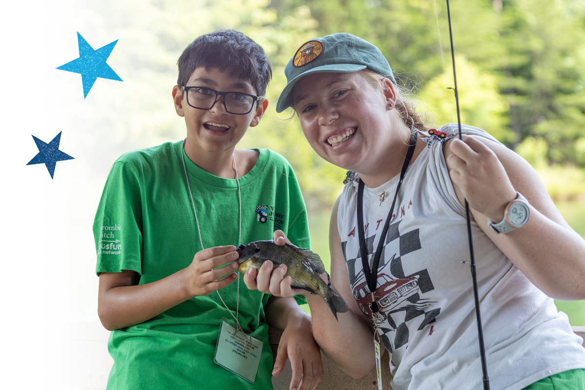Camper and counselor smile while holding a freshly caught fish by a lakeside, celebrating a shared outdoor experience.