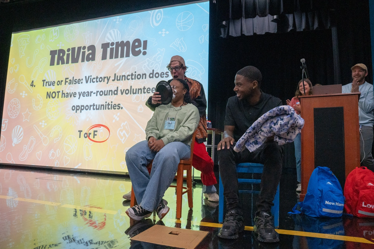Medical professionals participate in a camp-style trivia game on stage at Victory Junction during Med Camp 101, with one attendee playfully getting a pie to the face as others laugh and cheer.