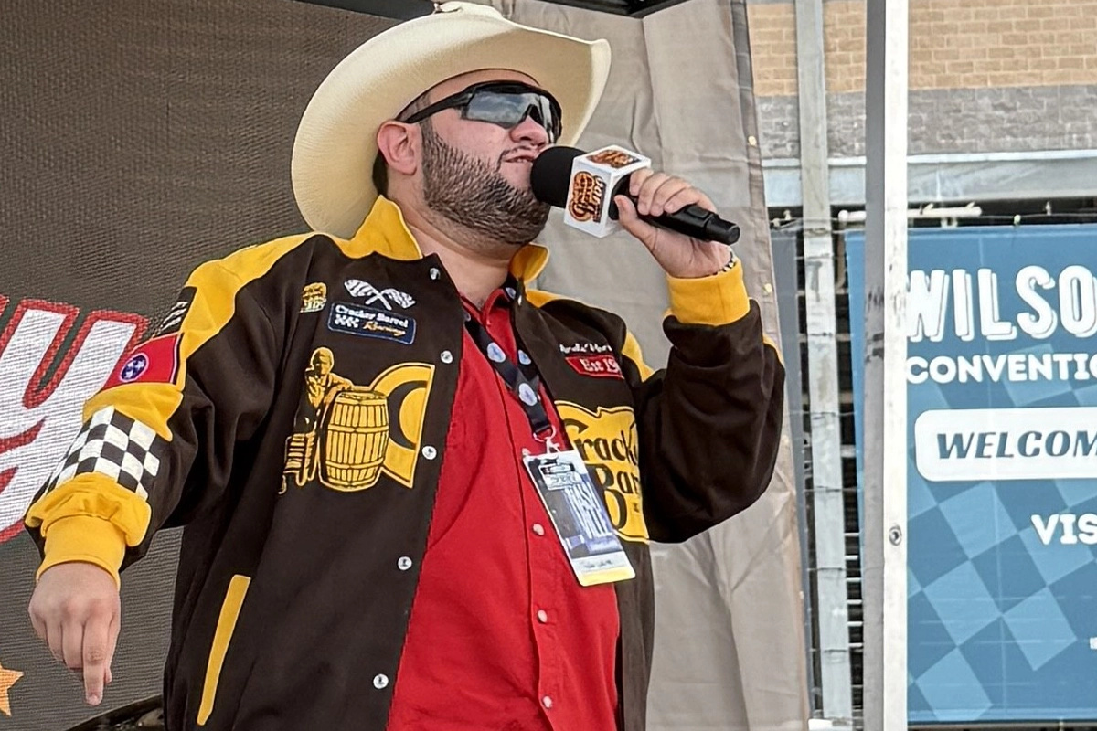 Country music artist Tyler White speaks into a microphone on an outdoor stage while wearing a white cowboy hat and a racing-themed jacket.