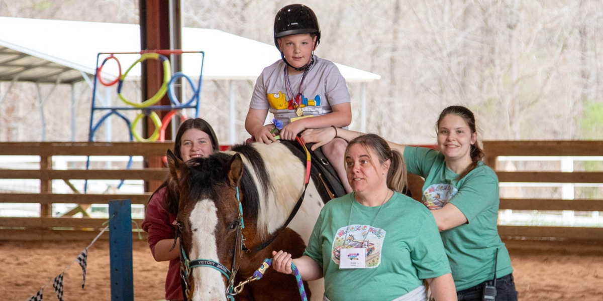 A camper wearing a helmet rides a paint horse during an adaptive horseback riding session while three volunteers walk alongside, guiding and supporting the experience in Victory Junction's covered riding arena.