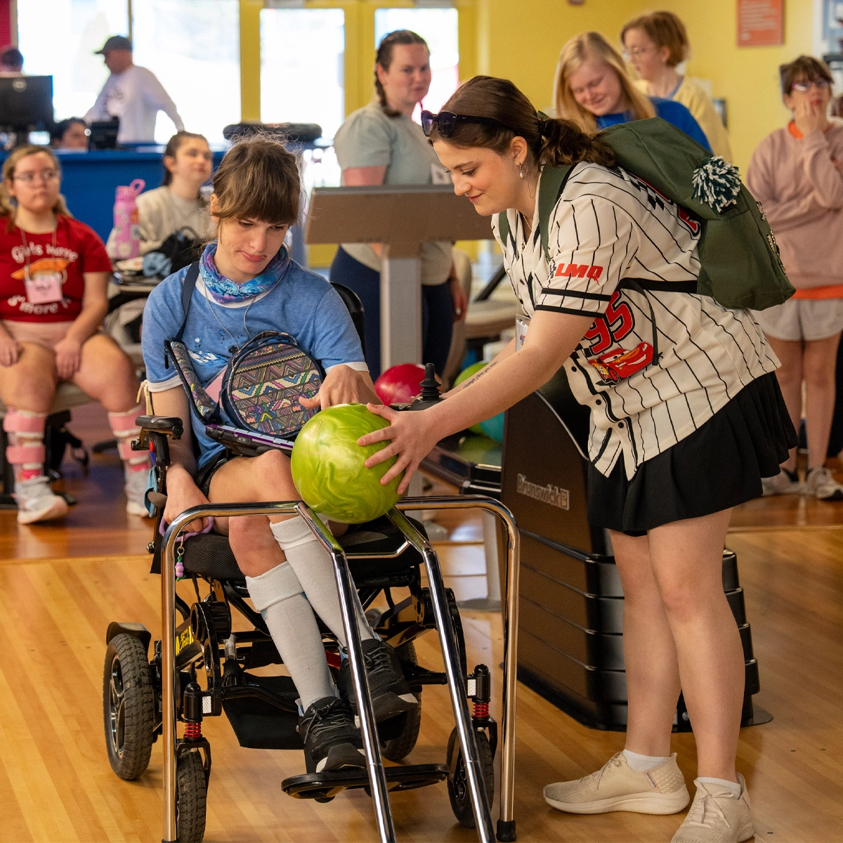 A camper using a power wheelchair rolls a bowling ball down an adaptive bowling ramp with assistance from a volunteer while other campers participate in the activity in the background.