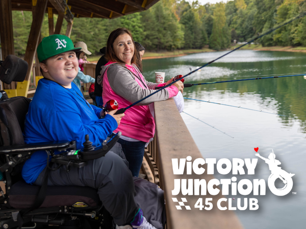 A young camper in a wheelchair and a parent or caregiver smile while fishing together from a covered dock overlooking a lake.
