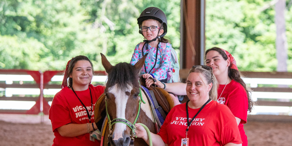 A young camper wearing a helmet rides a horse while three Victory Junction team members assist and offer support.