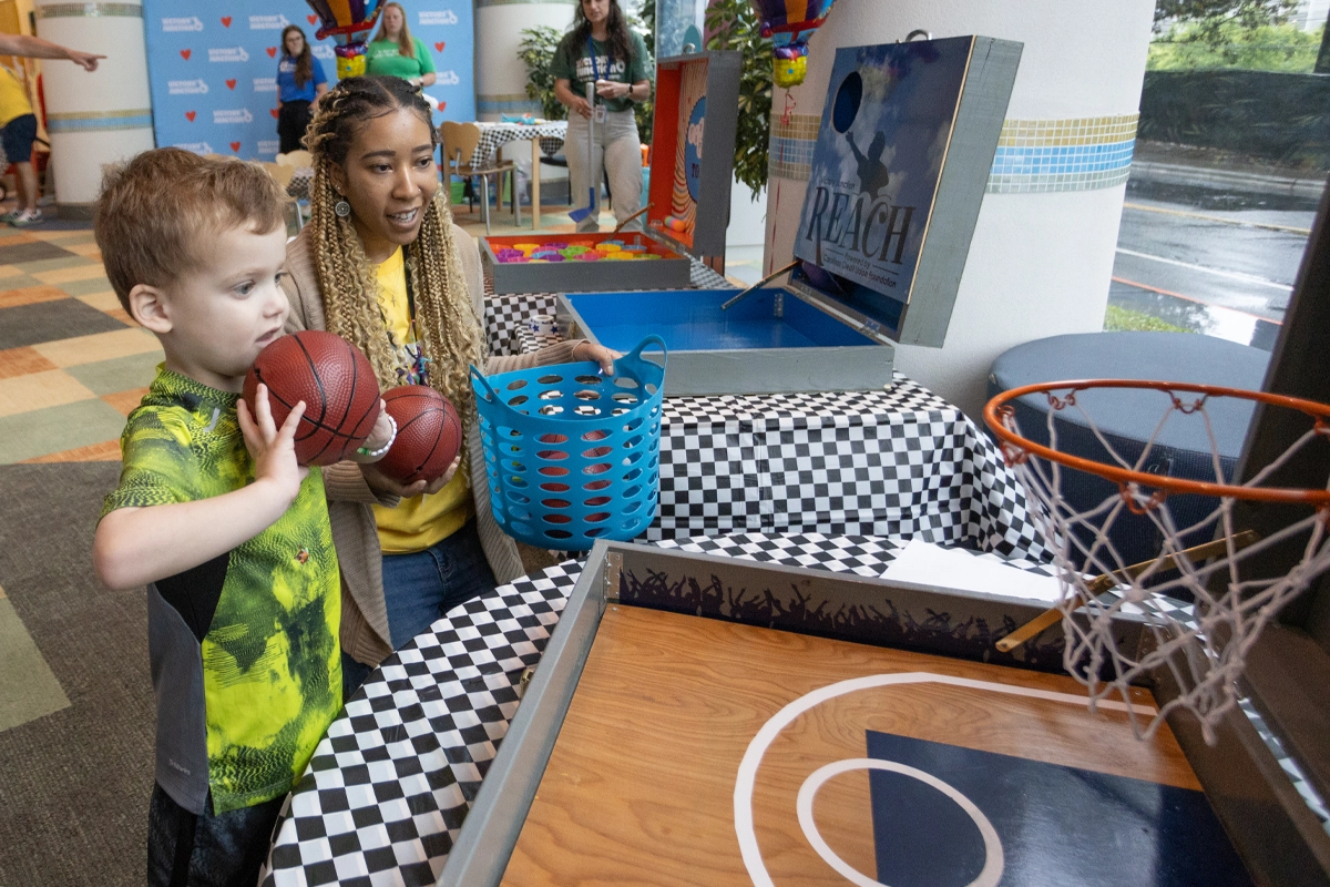 A young boy plays a basketball arcade game with encouragement from a Victory Junction staff member.
